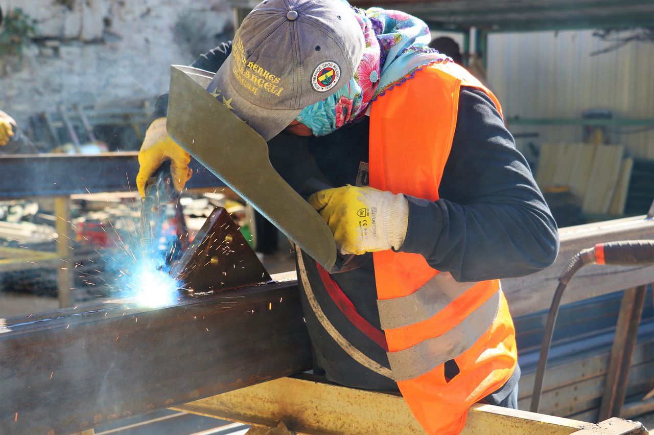 services-02 A welder wearing safety gear is welding metal beams, creating sparks outdoors.