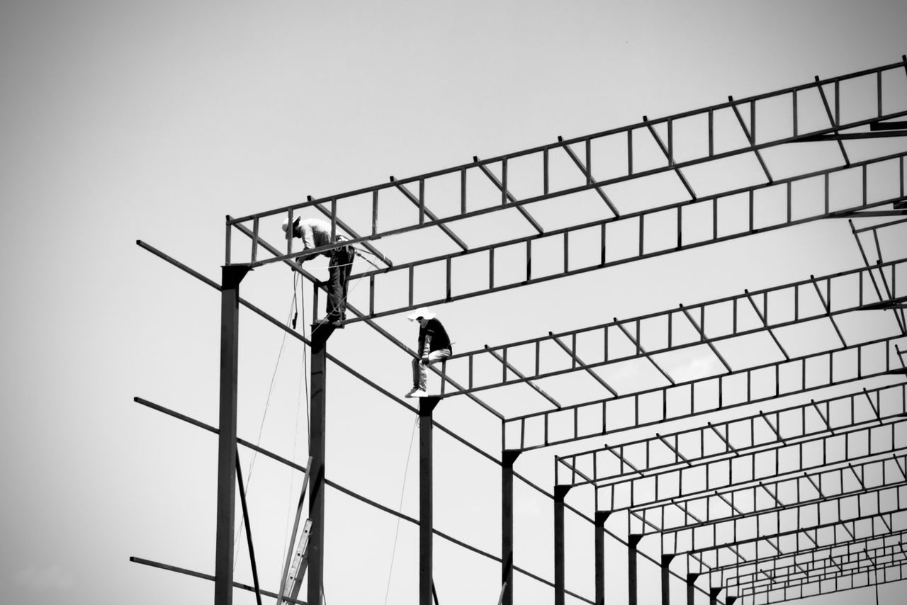 hero-img-01 Black and white shot of construction workers atop a steel frame structure under a clear sky.