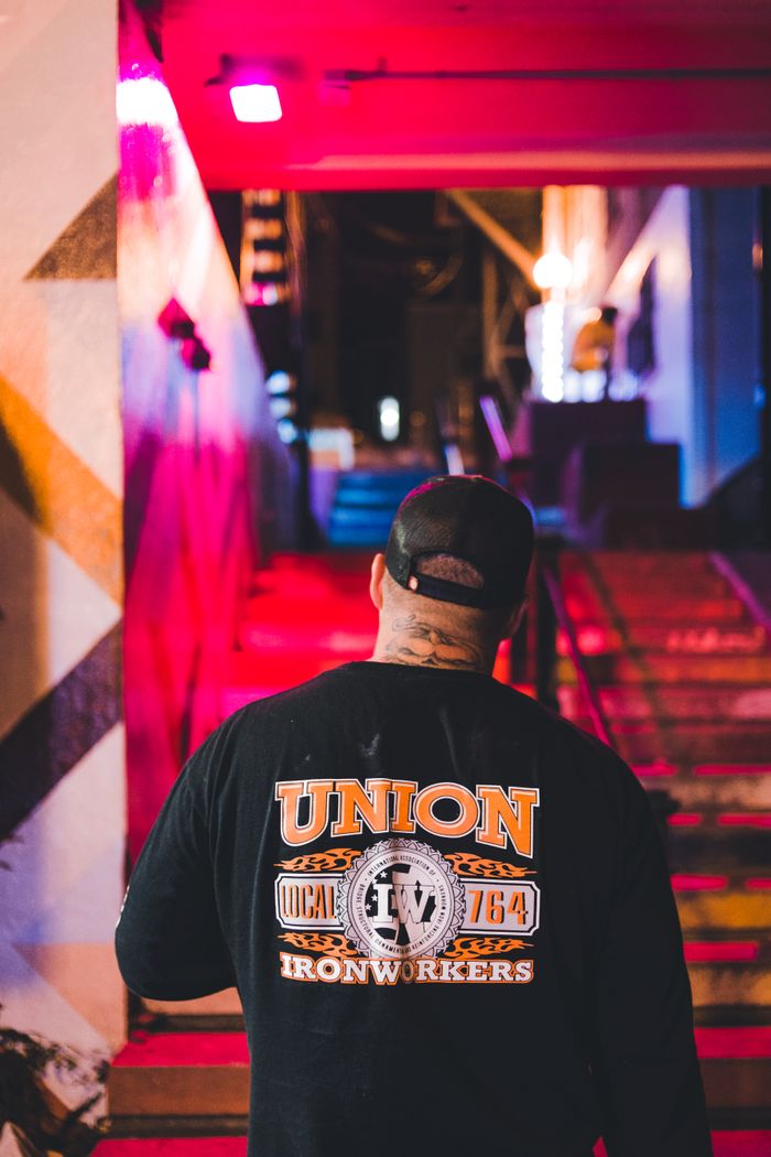 Man in ironworker union shirt walking up colorful urban stairs at night.